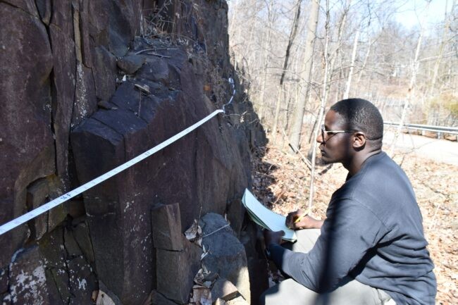 Man with notebook inspects cliff face closely.