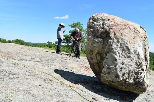 Scientists on rock face photographing a precariously balanced boulder.