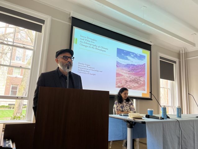 A man stands in front of a podium, with a panelists table and slideshow behind him.