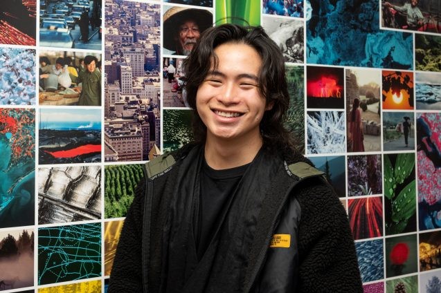 A smiling young man in front of a wall of photos