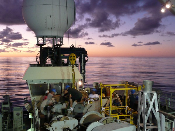 Crew from the R/V Atlantis work during the dawn hours to move a wire spool from one winch to another. The domed satellite dish that provides our Internet connection towers over the scene.