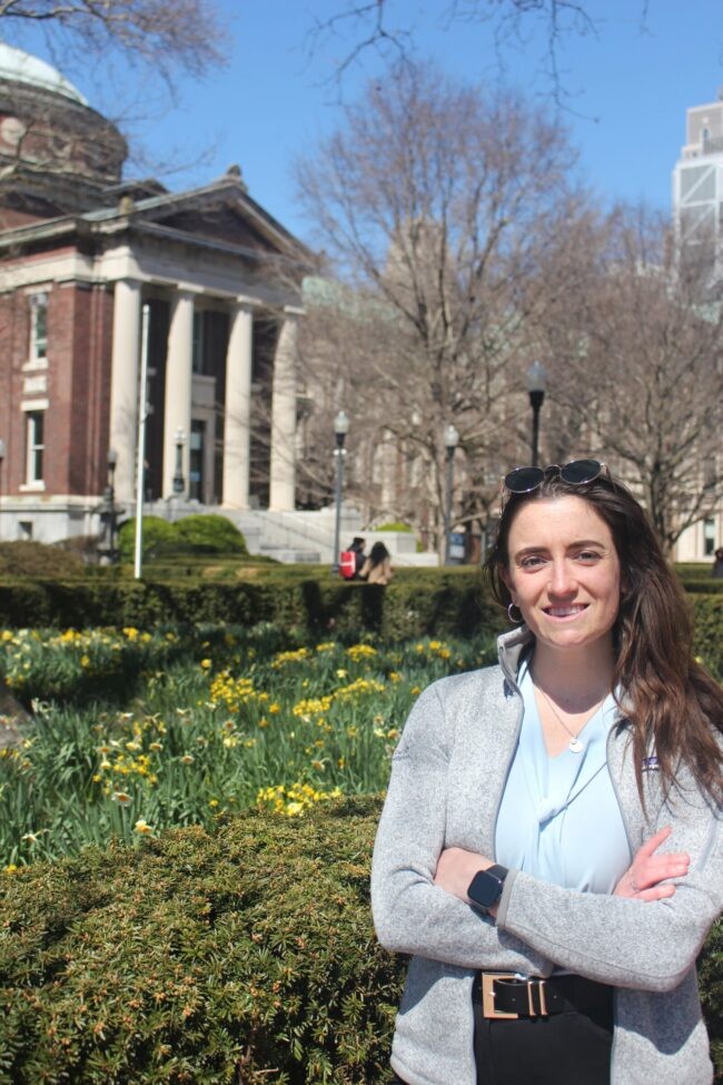 Woman stands with her arms crossed in front of building with columns and greenery