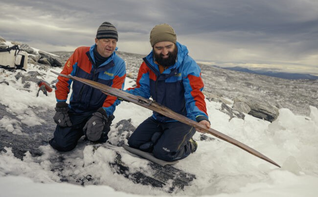 Two people in winter gear hold a wood ski on ice