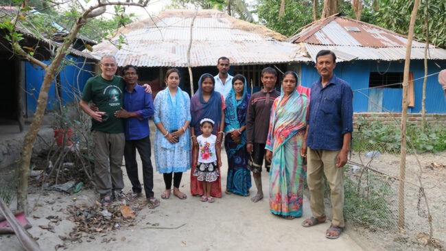 Family and visitors in Bangladesh pose in front of houses with thatched roofs