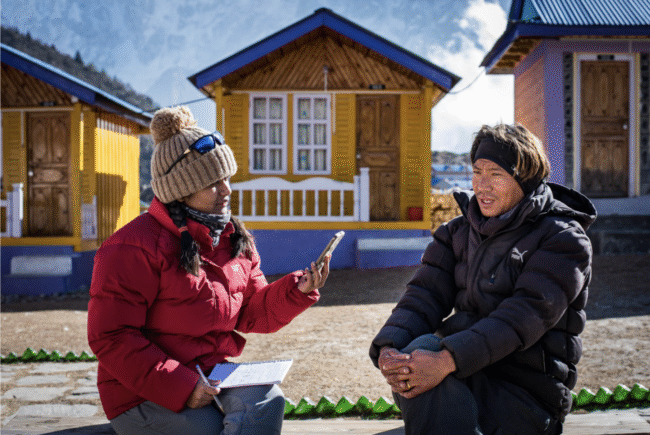 Woman in winter gear interviews man in front of a yellow and blue house.