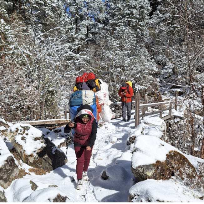 Female sherpa with bags on her back in front of a hiker on a snowy path