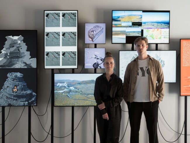 Two people stand in front of screens with wires against a wall