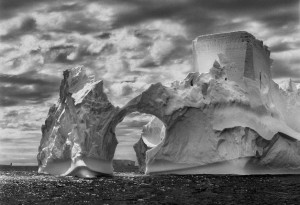 Sebastião Salgado, Iceberg between Paulet Island and the South Shetland Islands on the Antarctic Channel. At sea level, earlier flotation levels are clearly visible where the ice has been polished by the ocean’s constant movement. High above, a shape resembling a castle tower has been carved by wind erosion and detached pieces of ice. The Antarctic Peninsula, 2005. © Sebastião Salgado/Amazonas images—Contact Press Images.