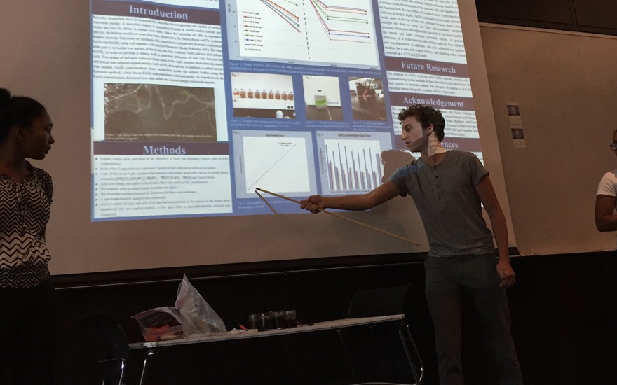 Josh Yurtsever describes how his team, working with Lamont's Ben Bostick, created a microbial battery from microbes in the marsh. The battery, on the desk in front of him, was able to light an LED lamp. SSRFP 2015 Presentations