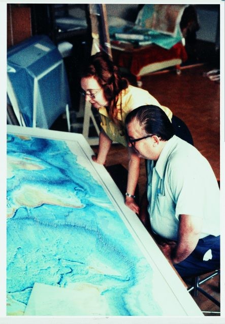Marie Tharp and Bruce Heezen sit in front of a world map of the ocean seafloor