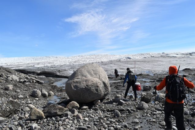 researchers walk across rocky terrain near edge of ice sheet