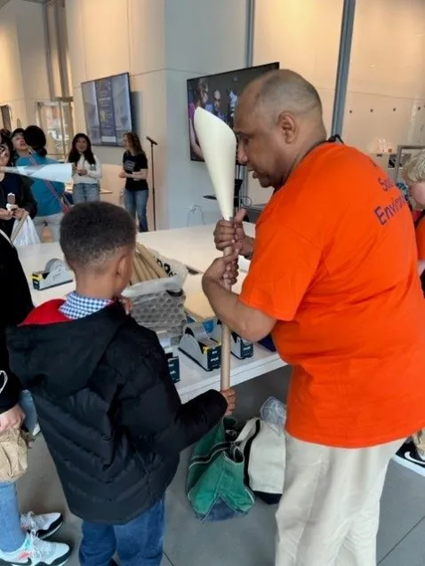 A man in an orane shirt helps a kid make an instrument from recycled materials