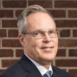 Man in glasses wearing a suit, against a brick backdrop.
