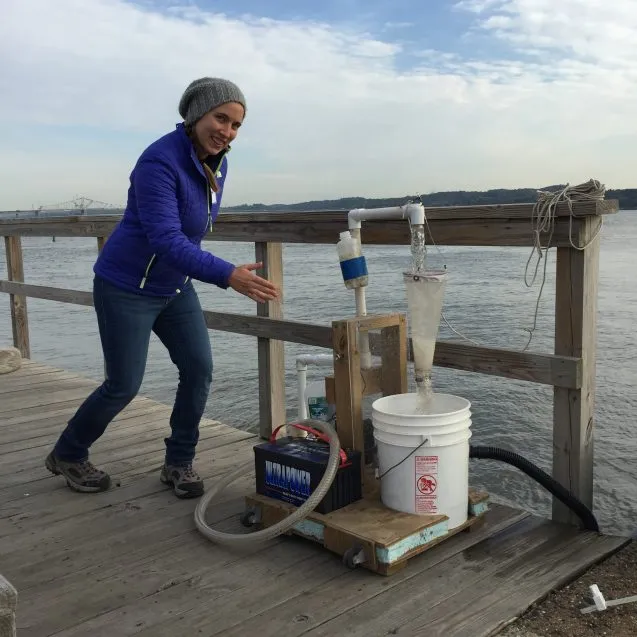 Woman in a hat points to pumping device near a river.