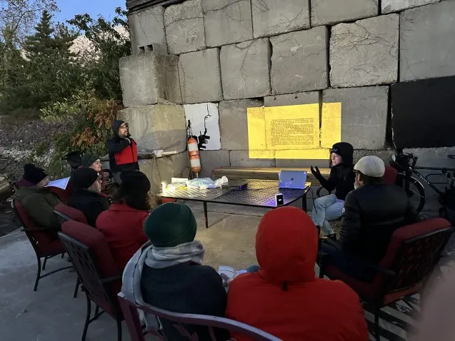 Group sits in a circle outside in winter clothing and looks at a presenter with projected text