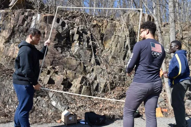 Two men carry a large frame made of plastic piping toward a cliff.