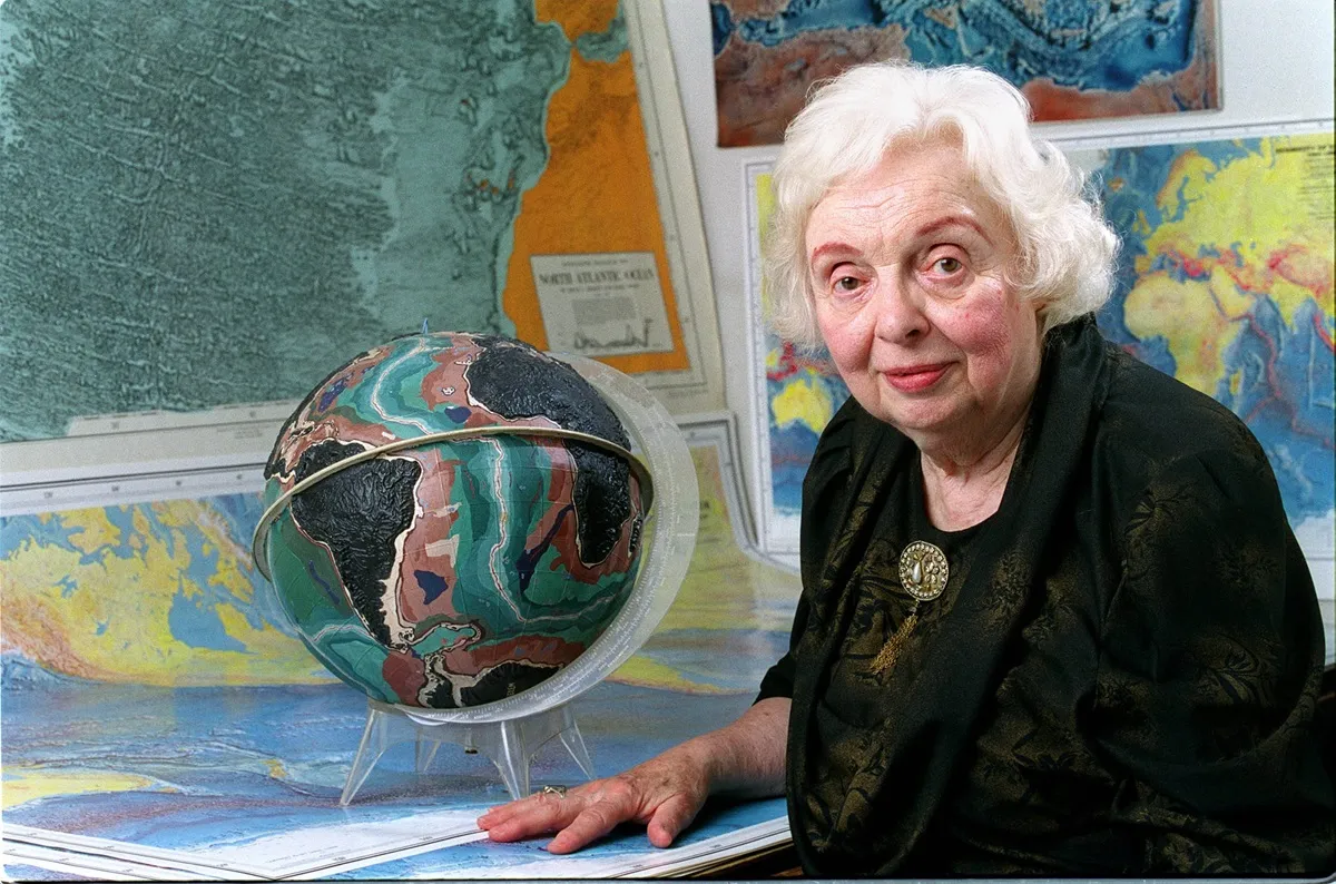 Marie Tharp in 2001 sitting in front of a bunch of maps and a model of the Earth