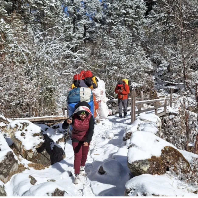 Female sherpa with bags on her back in front of a hiker on a snowy path