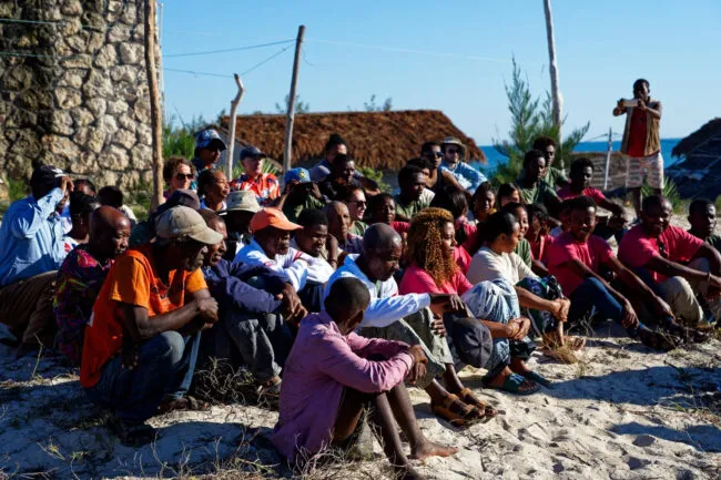 Photo of Douglass seated on sand among community members
