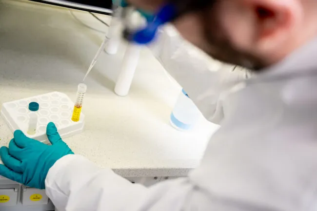 A scientist adds ammonia to a vial containing a yellow solution. The vial sits on a white lab bench. The researcher is wearing a whit lab coat and green gloves.