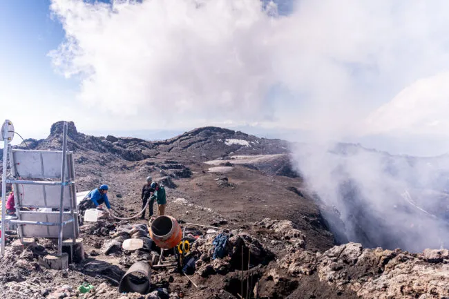 A gas plume from the volcano heads toward one of the instrument sites, where team members were working to set up the thermal and visual cameras. Solar panels (left)  were installed on steel frames. Photo by Francesco Fiondella.