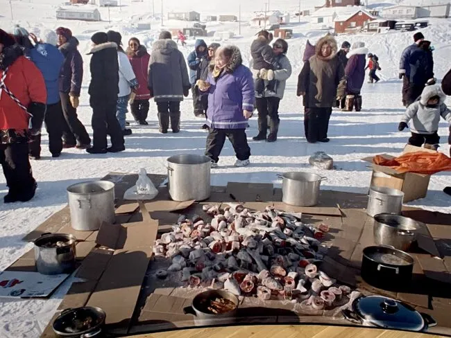 People gather around an outdoor fish preparation and cooking station in a snow-covered village