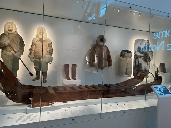 Museum exhibit displaying Inuit parkas, kayaks, and hunting equipment in a glass case