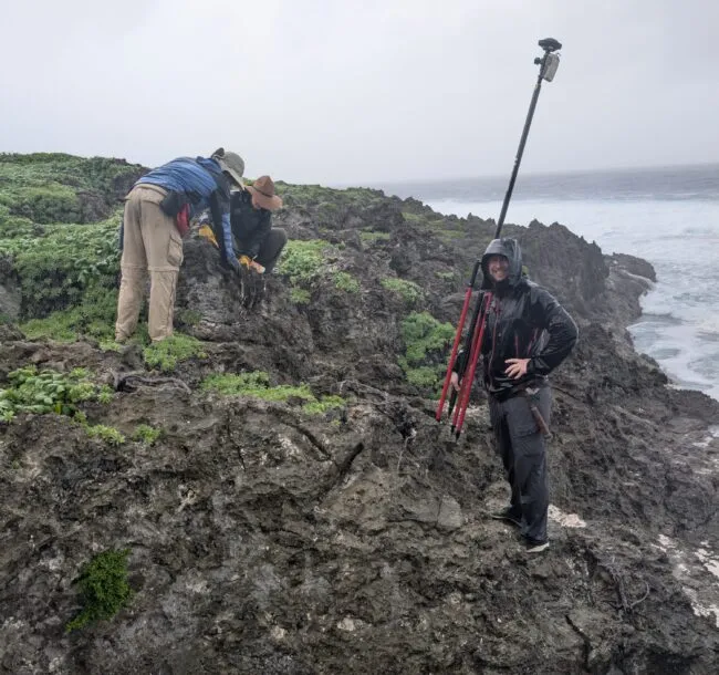 Three people in rain gear work on a rocky coastline beside the ocean, using surveying equipment