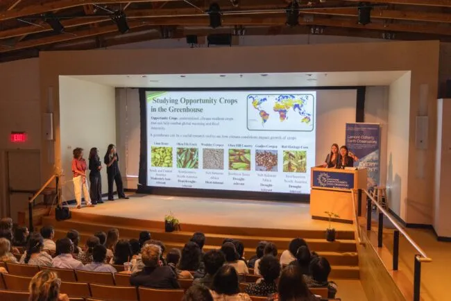 Three presenters discuss opportunity crops in a greenhouse as an audience views a slide on climate-resilient crops