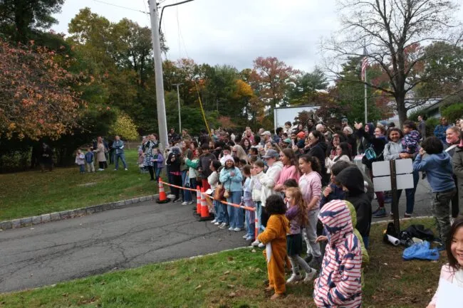 A large crowd of adults and children gathers outdoors in autumn, watching an event along a residential street