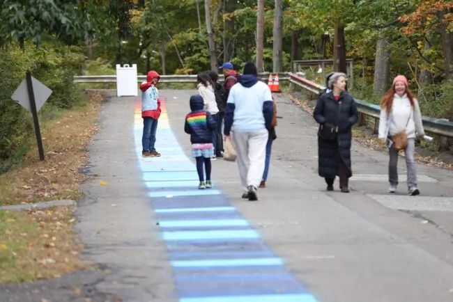 People walk along a wooded path marked with blue stripes, with a child in a red hood facing the camera