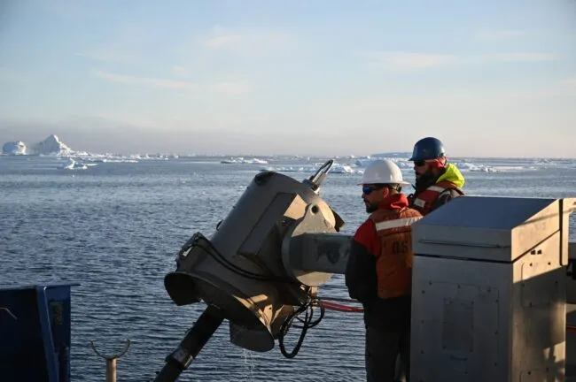 Two people lowering a jumbo piston corer into the sea. Ice dots the landscape behind them. 