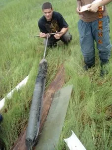 A student kneels in a grassy marsh, smiling with his hands on the handles of the large corer.